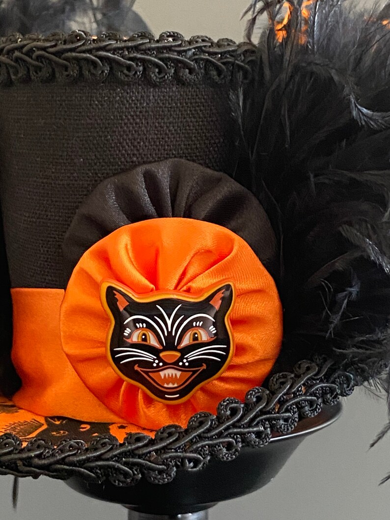 May include: A black top hat with an orange and black bow and a black cat face appliqu&eacute;. The hat is decorated with black feathers and black trim.