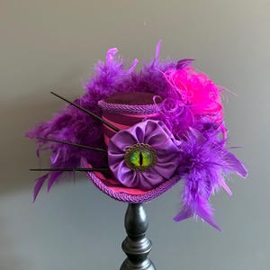 Puede incluir: Un sombrero de copa morado y rosa caprichoso con un adorno decorativo en forma de ojo. El sombrero est&aacute; adornado con plumas moradas, una flor de sat&eacute;n y detalles en negro. El sombrero se exhibe sobre un soporte negro.