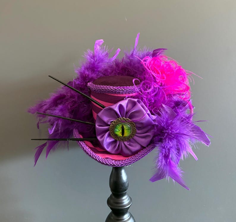 Puede incluir: Un sombrero de copa morado y rosa caprichoso con un adorno decorativo en forma de ojo. El sombrero est&aacute; adornado con plumas moradas, una flor de sat&eacute;n y detalles en negro. El sombrero se exhibe sobre un soporte negro.
