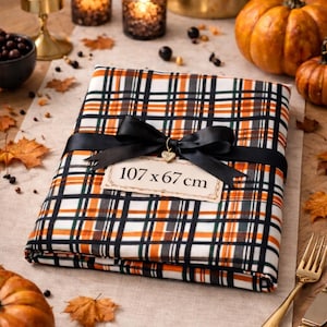 Peut inclure: Un tissu plié avec un ruban noir et une étiquette indiquant "107 x 67 cm". Le tissu présente un motif à carreaux orange, noir, blanc et vert. L'image a un thème automnal avec des citrouilles et des bougies.