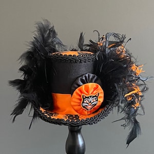 May include: A black top hat with an orange band and a black cat face appliqu&eacute;. The hat is decorated with black feathers.
