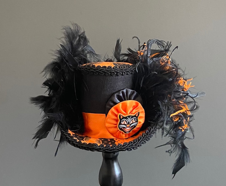 May include: A black top hat with an orange band and a black cat face appliqu&eacute;. The hat is decorated with black feathers.