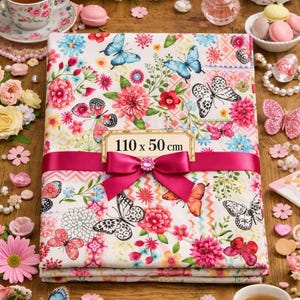 Peut inclure: Un tissu floral plié, mesurant 110 x 50 cm, est noué avec un ruban et un nœud roses. Le tissu présente un fond blanc avec des fleurs et des papillons colorés. L'image a un thème romantique et féminin.