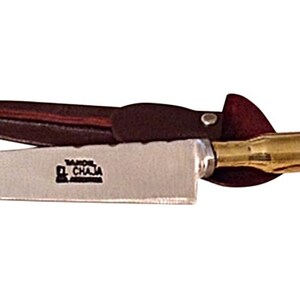 May include: A small, silver knife with a brass handle shaped like a bullet casing. The knife has a brown leather sheath with a metal snap closure. The blade is stamped with the words "Taller El Chala".