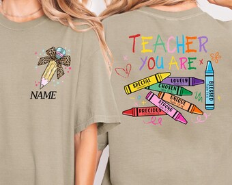 May include: A sage green t-shirt featuring a front graphic of a pencil with a leopard print bow and the word NAME. The back displays the text TEACHER YOU ARE with colorful crayons labeled SPECIAL, LOVELY, CHOSEN, UNIQUE, STRONG, PRECIOUS, and BLESSED.