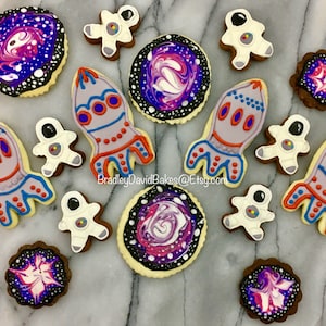 Puede incluir: Una colección de galletas de azúcar con temática espacial decoradas con glaseado morado, azul y naranja. Las galletas representan astronautas, cohetes y galaxias en espiral.