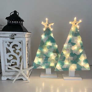 May include: Two teal blue and white mosaic Christmas trees with white starfish on top and white bases. The trees are lit with warm white lights.