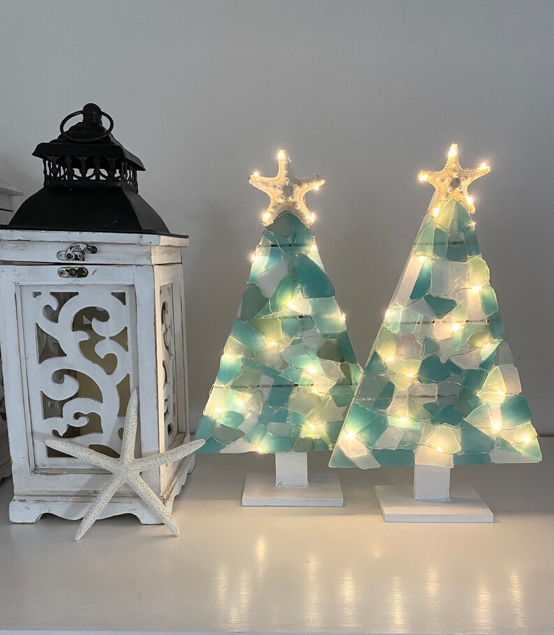 May include: Two teal blue and white mosaic Christmas trees with white starfish on top and white bases. The trees are lit with warm white lights.