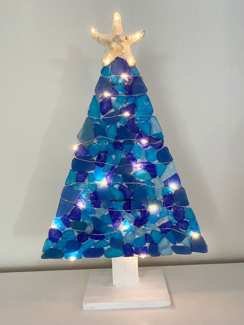 May include: A blue and white sea glass Christmas tree with white lights and a starfish on top. The tree is on a white wooden base.