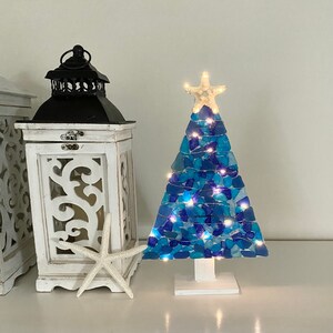 May include: A blue and white sea glass Christmas tree with white lights. The tree is decorated with a white starfish on top and is sitting on a white base.