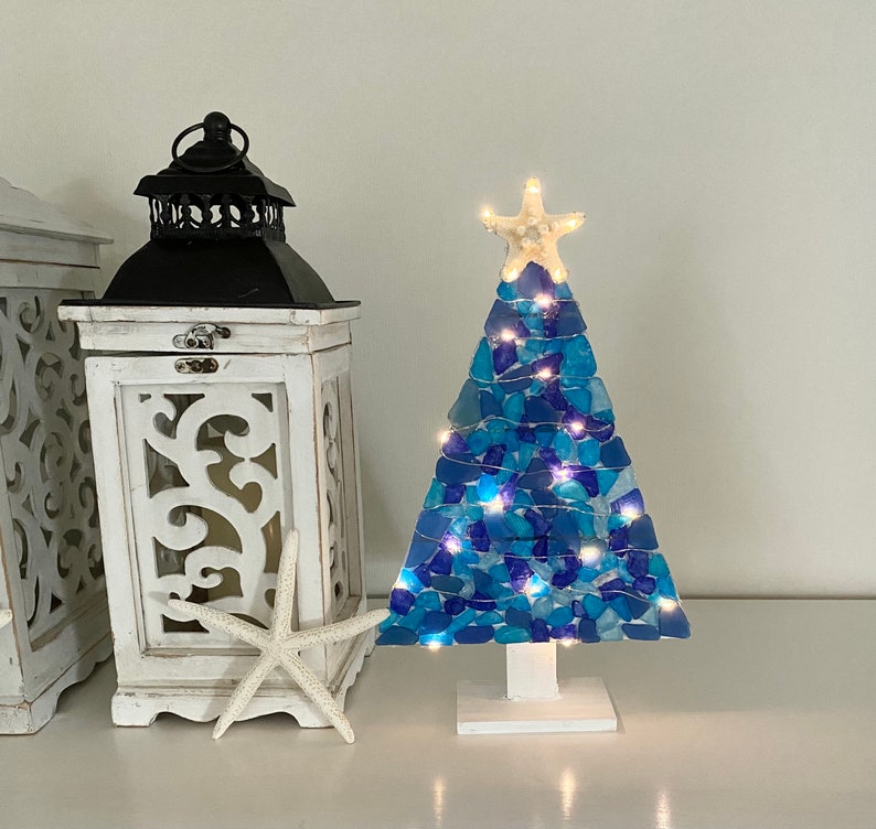 May include: A blue and white sea glass Christmas tree with white lights. The tree is decorated with a white starfish on top and is sitting on a white base.