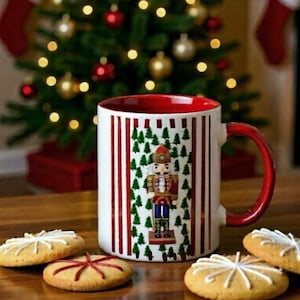 Può includere: Una tazza di ceramica bianca con interno e manico rossi, decorata con un disegno di schiaccianoci e strisce rosse verticali. Biscotti decorati e un albero di Natale sullo sfondo.