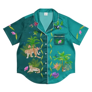 以下が含まれることがあります： ターコイズブルーの半袖ボタンダウンシャツ。トロピカルなプリントが施され、ヒョウ、ヤシの木、花、蝶が描かれています。