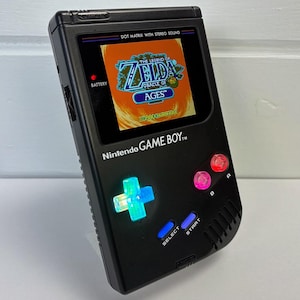 May include: Black Nintendo Game Boy handheld console with a screen displaying "The Legend of Zelda: Oracle of Ages." The console has illuminated buttons and the text "DOT MATRIX WITH STEREO SOUND" and "Nintendo GAME BOY."