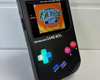 May include: Black Nintendo Game Boy handheld console with a screen displaying "The Legend of Zelda: Oracle of Ages." The console has illuminated buttons and the text "DOT MATRIX WITH STEREO SOUND" and "Nintendo GAME BOY."