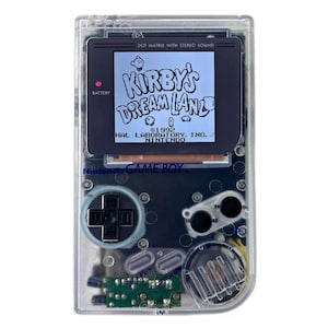 May include: A clear, transparent Game Boy Color handheld video game console with a black and white screen displaying the title "Kirby's Dream Land". The console has a black D-pad, two black buttons, and a black start button.