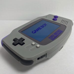 May include: A gray Game Boy Advance SP handheld gaming console with a purple trim. The screen displays the text "GAME BOY" and "SUPER NINTENDO" in blue. The console has a black directional pad, white start and select buttons, and a purple power button.