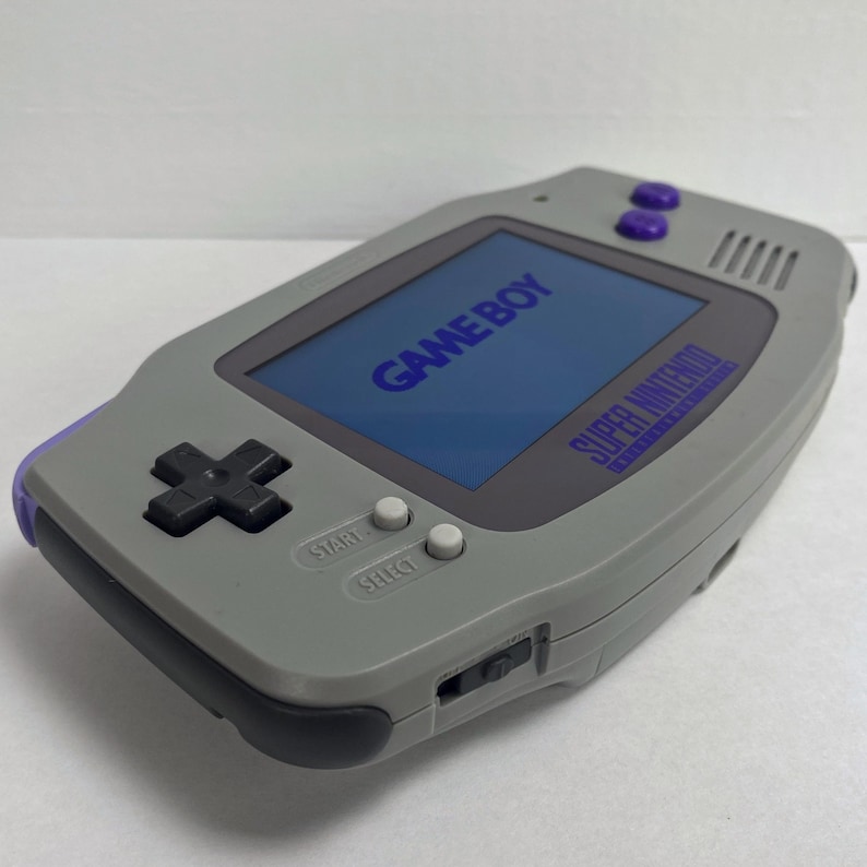 May include: A gray Game Boy Advance SP handheld gaming console with a purple trim. The screen displays the text "GAME BOY" and "SUPER NINTENDO" in blue. The console has a black directional pad, white start and select buttons, and a purple power button.