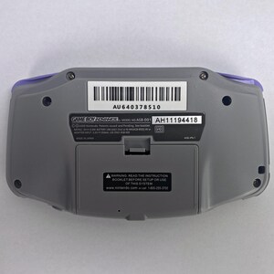 May include: A gray Game Boy Advance handheld gaming console with a purple trim. The back of the console has a sticker with the model number, serial number, and warning label. The text on the sticker reads: "GAME BOY ADVANCE MODEL NO. AGB-001 AU640378510 AH11194418 2000 Nintendo Patents issued and Pending. See booklet. RATING: 3.3V DC BATTERY: LR6 (AA) x 2 (MADE IN JAPAN) ADAPTER INPUT: 3.3V DC USE ONLY AGB-108 MADE IN JAPAN A-1 WARNING: READ THE INSTRUCTION BOOKLET BEFORE SETUP OR USE OF THIS SYSTEM www.nintendo.com or call 1-800-255-3700"