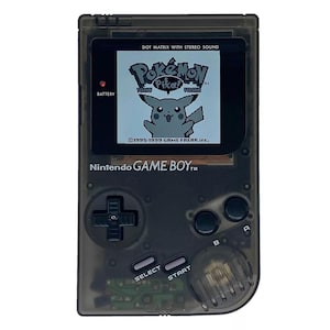 May include: A black Game Boy Color handheld video game console with a translucent shell. The screen displays a Pokemon game with Pikachu on the screen. The text on the screen reads "Pokemon Pika! Version 1995-1999 GAME FREAK inc."