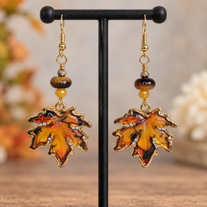 May include: A pair of gold-tone dangle earrings featuring amber-colored maple leaf charms with small clear crystals. Each earring includes a round brown stone bead, a small yellow bead, and a gold-tone hook wire closure.
