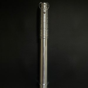 May include: A silver lightsaber hilt with a cylindrical design. The hilt features a series of rings and grooves, with a circular emitter at the top. The bottom has a small, rounded base. The lightsaber is made of metal and has a polished finish.