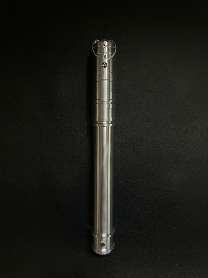 May include: A silver lightsaber hilt with a cylindrical design. The hilt features a series of rings and grooves, with a circular emitter at the top. The bottom has a small, rounded base. The lightsaber is made of metal and has a polished finish.