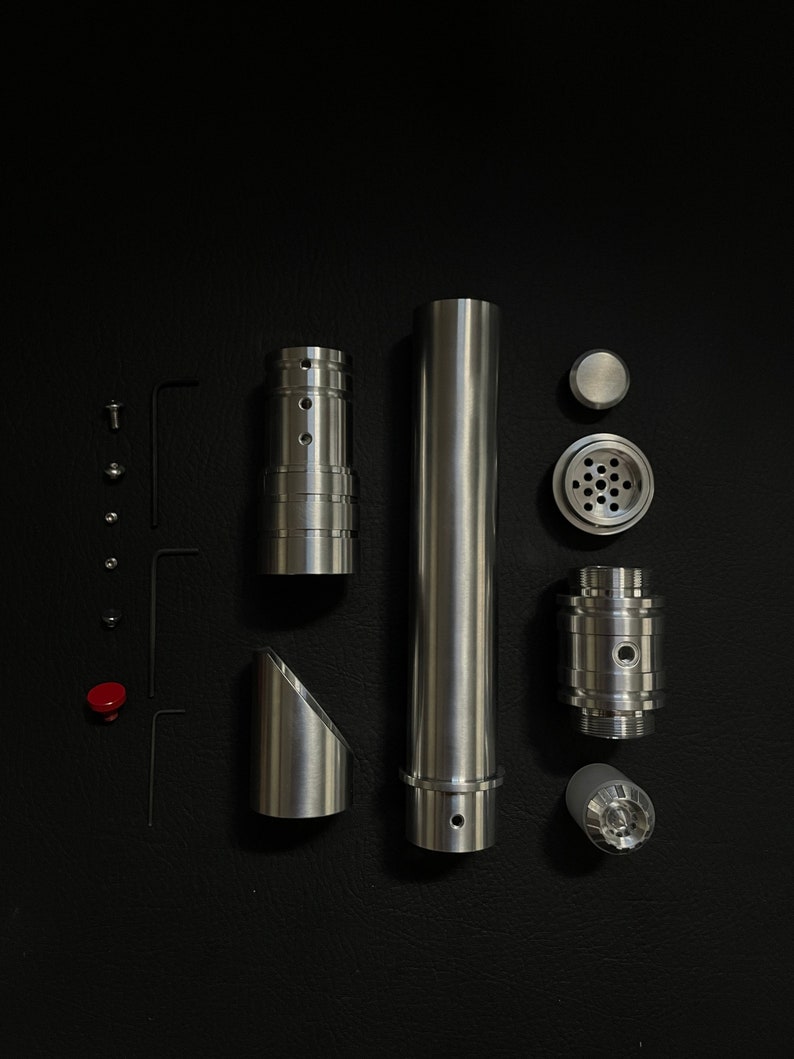 May include: An assortment of disassembled metallic components, including cylindrical tubes, angled pieces, and smaller parts like screws and a red button, arranged on a black surface. The pieces have a brushed metal finish.