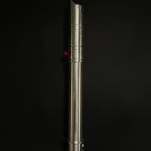 May include: A silver lightsaber with a cylindrical handle and a beveled emitter. The handle features ridged sections and a red button. The lightsaber is made of metal and has a polished finish. The background is black.