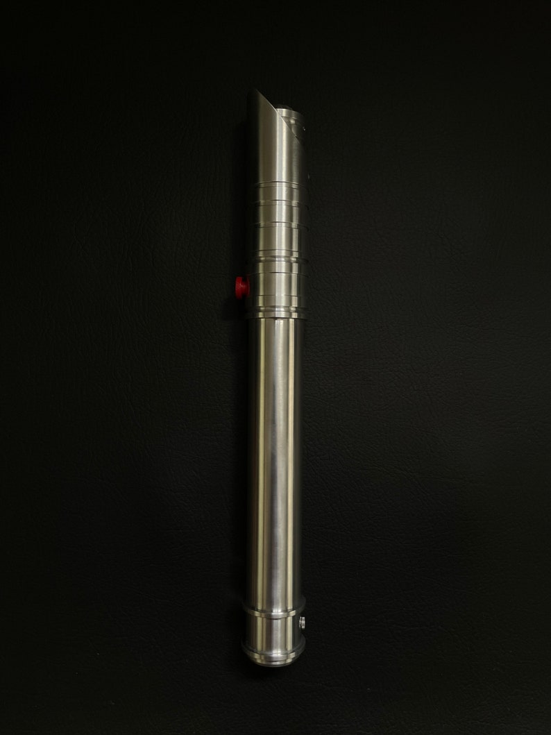 May include: A silver lightsaber with a cylindrical handle and a beveled emitter. The handle features ridged sections and a red button. The lightsaber is made of metal and has a polished finish. The background is black.