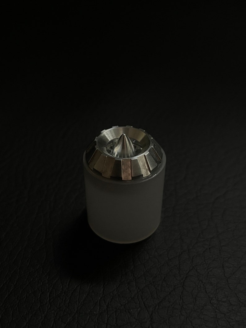 May include: A cylindrical, frosted glass object with a metallic, conical top. The top has a series of rectangular, silver-colored segments around the base of the cone. The object is set against a dark, textured background.