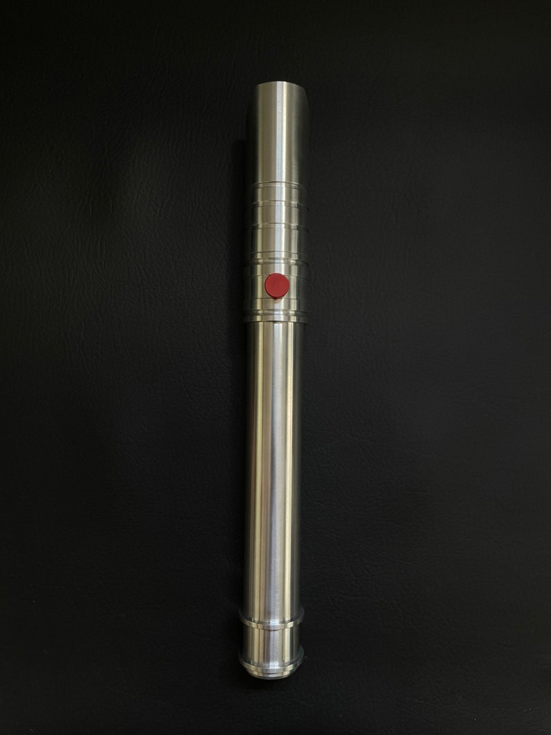 May include: A silver-colored lightsaber with a red button. The hilt has a cylindrical design with ridged sections and a smooth, polished finish. The lightsaber is a prop or toy, likely for cosplay or display.