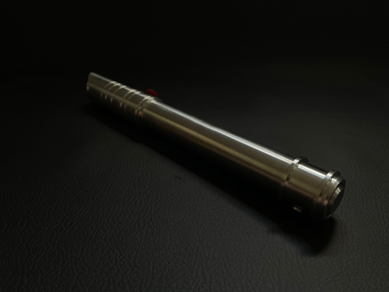May include: A metallic silver lightsaber prop with a red button, lying on a black surface. The lightsaber has a cylindrical body with a ridged grip section and a detailed emitter.