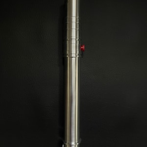 May include: A silver lightsaber with a brushed metal finish. The hilt features a red button and a series of circular ridges. The blade emitter has a curved design. The lightsaber is displayed against a black background.