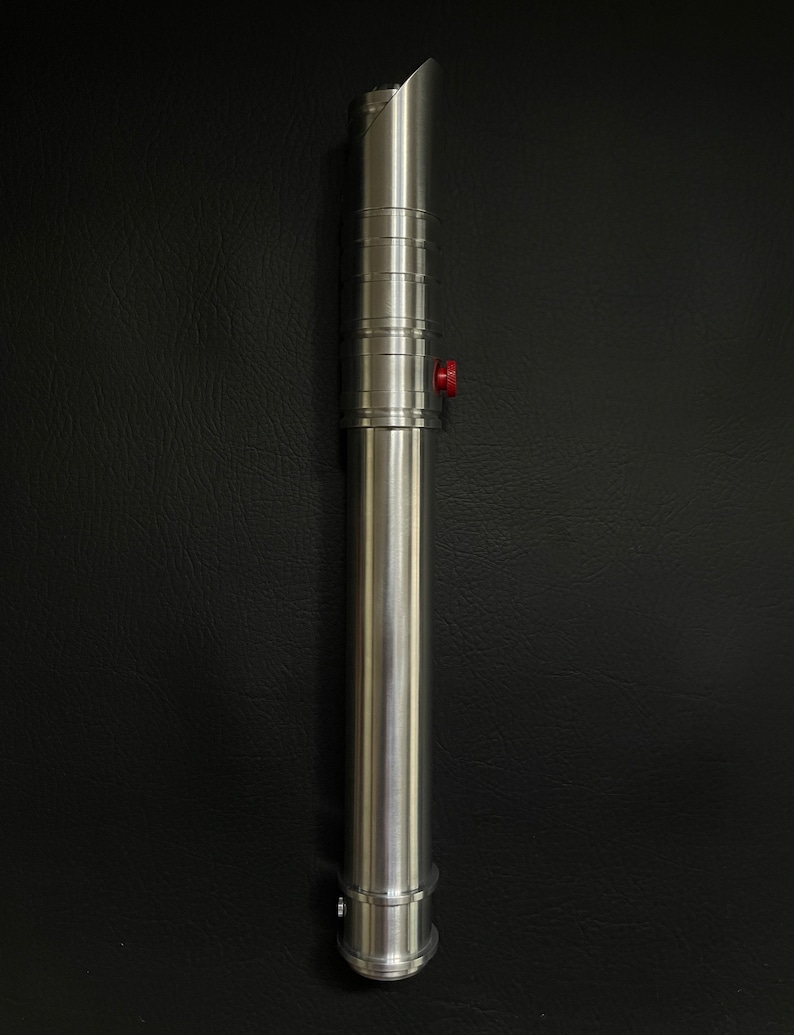 May include: A silver lightsaber with a brushed metal finish. The hilt features a red button and a series of circular ridges. The blade emitter has a curved design. The lightsaber is displayed against a black background.