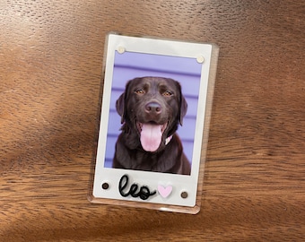 May include: A clear acrylic photo holder with a picture of a brown Labrador Retriever dog with its tongue out. The photo has a white border and the name "leo" in black script with a pink heart.