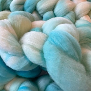 May include: A close-up of a pile of soft, fluffy yarn in shades of white and light blue. The yarn is arranged in a braided pattern, creating a textured and visually appealing display.