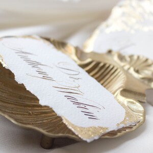 K&ouml;nnte beinhalten: Goldfarbener Muschelschalentischkartenhalter mit einer wei&szlig;en Papierkarte, auf der "Dr. Martin" in eleganter Kalligraphie steht.