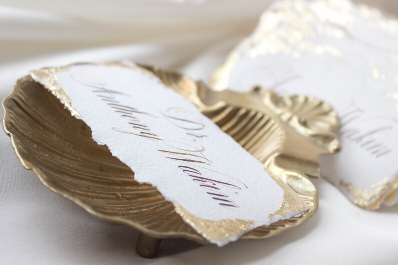 K&ouml;nnte beinhalten: Goldfarbener Muschelschalentischkartenhalter mit einer wei&szlig;en Papierkarte, auf der "Dr. Martin" in eleganter Kalligraphie steht.
