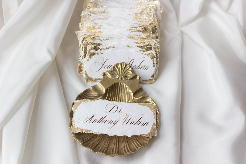 K&ouml;nnte beinhalten: Eine goldfarbene, muschelf&ouml;rmige Schale mit einer wei&szlig;en Karte darin. Die Karte hat goldene Blattverzierungen und den Text "Dr. Anthony Wakim" in brauner Tinte.