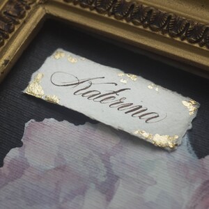 K&ouml;nnte beinhalten: Eine wei&szlig;e Papierkarte mit Blattgoldakzenten und dem Wort "Aterna" in Kalligraphie geschrieben.
