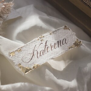 K&ouml;nnte beinhalten: Eine wei&szlig;e Papier-Platzkarte mit goldenen Akzenten und dem Namen "Katerina" in Kalligraphie.