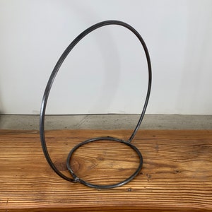 May include: A metal stand with a circular base and a large, curved loop extending upwards. The stand is made of a dark, metallic material and has a simple, minimalist design.