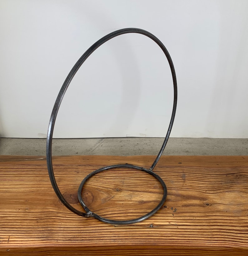 May include: A metal stand with a circular base and a large, curved loop extending upwards. The stand is made of a dark, metallic material and has a simple, minimalist design.
