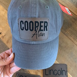 May include: Three brown leather patches with black embroidered text. The patches read "Beau" with an arrow, "Lincoln", and "Cooper Alan". A gray baseball cap with the "Cooper Alan" patch attached is also shown.
