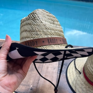May include: A straw cowboy hat with a black and white chequered brim. The hat has a brown band with the name "BRODY" printed on it. The hat is held by a person's hand.