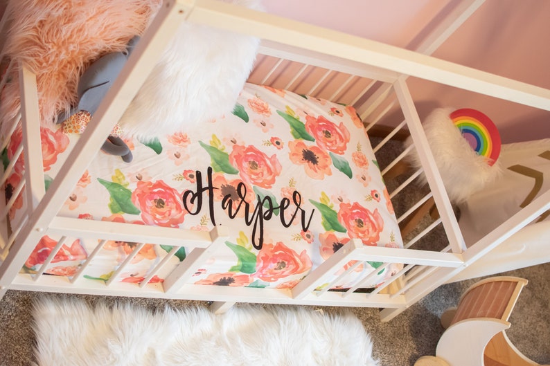 May include: A white wooden bed frame with a floral patterned bed sheet. The sheet features large coral and peach flowers with green leaves and the name "Harper" in black script. A fluffy white rug and pillows are also visible.