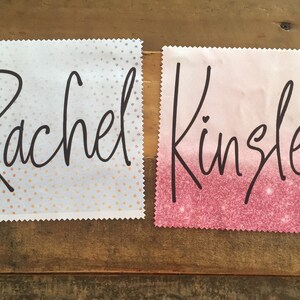 Op de afbeelding: Twee witte vierkante stoffen met roze en witte stippen en roze glitterpatronen. De namen "Rachel" en "Kingsley" zijn in zwarte cursieve letters op elk vierkant geschreven.