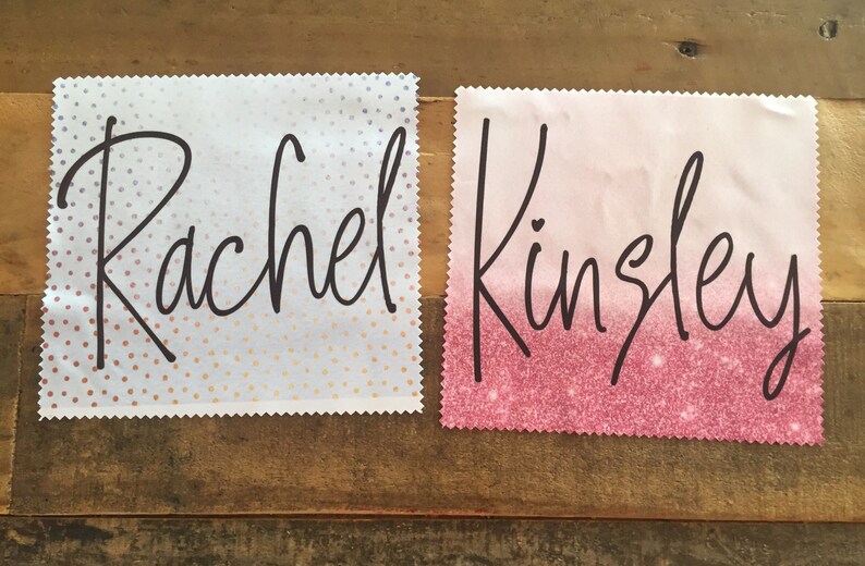 Op de afbeelding: Twee witte vierkante stoffen met roze en witte stippen en roze glitterpatronen. De namen "Rachel" en "Kingsley" zijn in zwarte cursieve letters op elk vierkant geschreven.