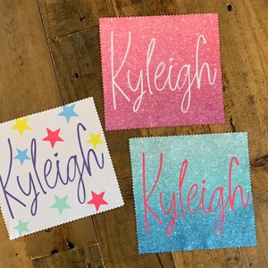 Op de afbeelding: Drie gepersonaliseerde stoffen vierkanten met de naam "Kyleigh" geschreven in verschillende stijlen. Het eerste vierkant heeft een witte achtergrond met kleurrijke sterren. Het tweede vierkant heeft een roze glitterachtergrond. Het derde vierkant heeft een blauwe en groene ombre glitterachtergrond.